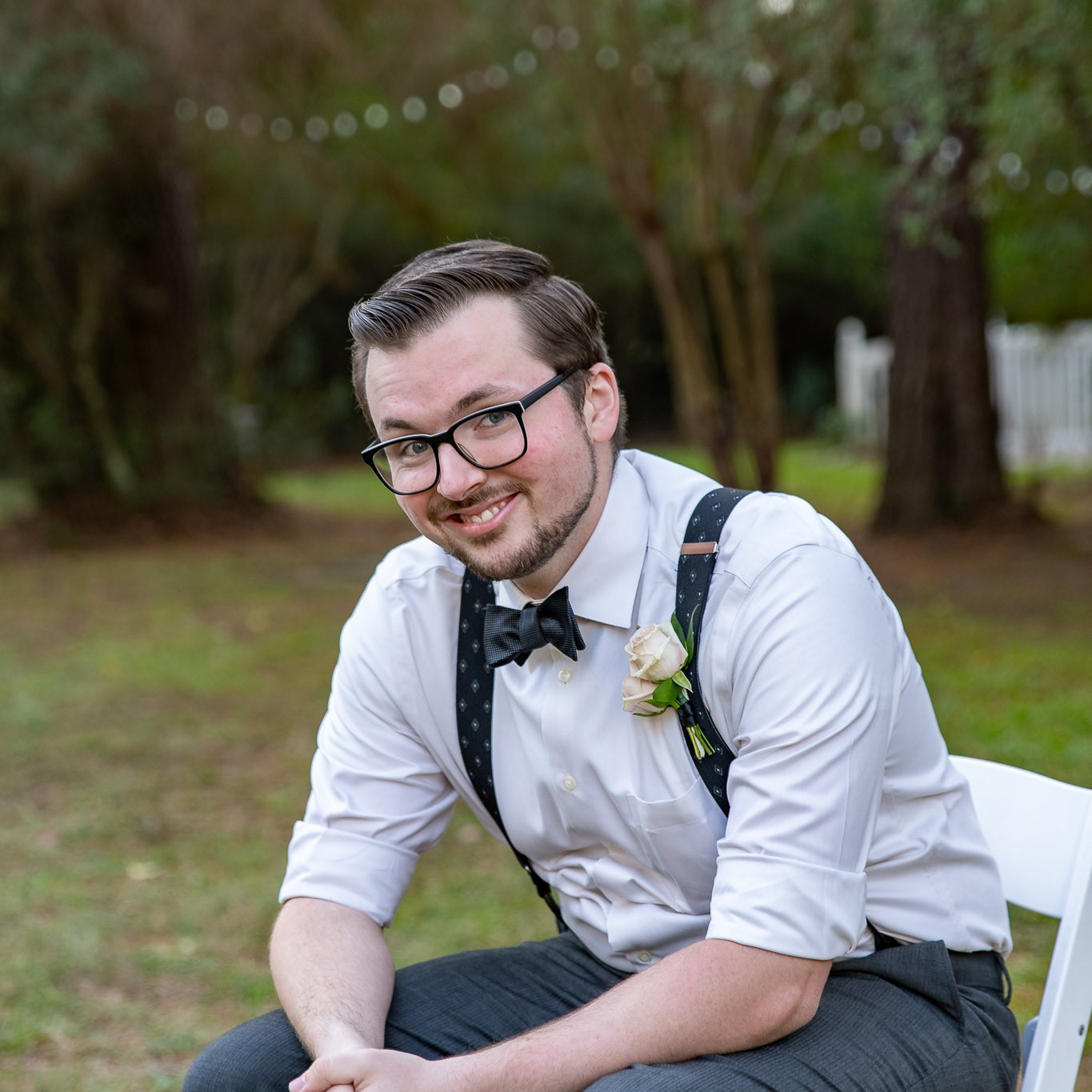 A photo of me! I'm seated outdoors on a white chair in mostly-formal dress, clad in gray trousers, a white french-cuffed shirt rolled up to the elbows, black suspenders, a black bow tie, and my usual crisp pompadour haircut. I'm beaming-- a huge grin is hardly hidden by my short beard and mustache, and my eyes smile out from behind huge black-framed glasses. A white carnation clings to one suspender strap.